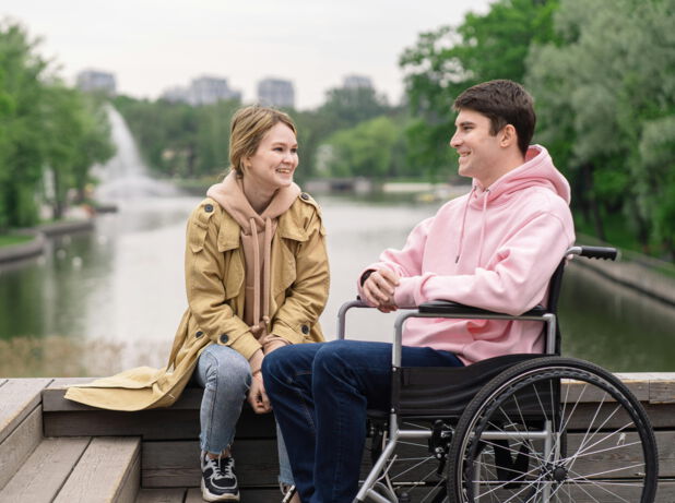 Das Bild zeigt einen  jungen Mann, der in einem Rollstuhl sitzt, auf einer Brücke. Sein Gesicht ist einer jungen Frau zugewandt, die links von ihm auf einer Mauer sitzt. Sein Mund ist leicht geöffnet. Sie lächelt ihn an. Im Hintergrund befinden sich die Wasseroberfläche eines Flusses und die grünbelaubten Bäume eines Parks.