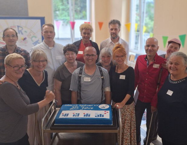 Eine Gruppe von 13 Menschen steht in einem hell erleuchteten Raum zusammen. Alle blicken uns an. Vor ihnen steht ein rechteckiger, blau glasierter Kuchen auf einem Servierwagen. Auf dem Kuchen steht in weißer Schrift: „Leben mit Handicaps e.V. – 25 Jahre“. Ebenfalls befinden sich blaue Punkte und Kreise auf dem Kuchen, die an das Logo des Vereins erinnern. Im Hintergrund hängen bunte Wimpelgirlanden und ein Plakat mit Zeichnungen. Alle Personen wirken fröhlich und lächeln.